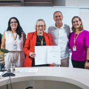 A imagem mostra um grupo de cinco pessoas reunido atr&aacute;s de uma mesa semicircular em ambiente institucional. A pessoa ao centro segura um documento aberto, exibido voltado para a c&acirc;mera.