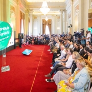 Sal&atilde;o amplo e elegante, com tapete vermelho e lustres grandes. &Agrave; esquerda, Leite fala ao microfone diante de um tel&atilde;o verde com a frase &ldquo;O Rio Grande t&aacute; diferente &ndash; Parcerias em a&ccedil;&atilde;o&rdquo;. &Agrave; direita, uma plateia numerosa sentada observa, enquanto c&acirc;meras de TV registram o evento.