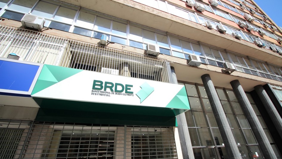 Vis&atilde;o Geral
A imagem mostra a fachada de um edif&iacute;cio comercial de m&uacute;ltiplos andares, com foco em uma placa de identifica&ccedil;&atilde;o do BRDE (Banco Regional de Desenvolvimento do Extremo Sul). A foto &eacute; tirada de um &acirc;ngulo de baixo para cima (contra-plong&eacute;e), enfatizando a altura da estrutura.

Detalhes da Fachada e Identifica&ccedil;&atilde;o
Placa do Banco: Em primeiro plano, no lado esquerdo, h&aacute; um painel horizontal moderno com fundo branco e detalhes geom&eacute;tricos em tons de verde. Nele, l&ecirc;-se em letras escuras: "BRDE - BANCO REGIONAL DE DESENVOLVIMENTO DO EXTREMO SUL", acompanhado por um logotipo verde que remete ao mapa da regi&atilde;o Sul do Brasil.

O Edif&iacute;cio: A estrutura possui uma arquitetura funcional, com janelas de vidro que refletem a luz do dia e diversas unidades de ar-condicionado fixadas na parede externa de tijolos aparentes. Colunas cil&iacute;ndricas escuras sustentam a parte frontal do pr&eacute;dio no n&iacute;vel da cal&ccedil;ada.

Entrada: Abaixo da placa do BRDE, v&ecirc;-se uma grade met&aacute;lica de prote&ccedil;&atilde;o e uma porta de vidro que d&aacute; acesso ao interior do banco.

Composi&ccedil;&atilde;o e Ilumina&ccedil;&atilde;o
Perspectiva: O &acirc;ngulo acentuado cria uma linha diagonal que guia o olhar da placa no canto inferior esquerdo at&eacute; o topo do edif&iacute;cio no canto superior direito.

Ilumina&ccedil;&atilde;o: A luz solar &eacute; direta e clara, indicando um dia ensolarado. O c&eacute;u azul &eacute; vis&iacute;vel no canto superior direito, contrastando com os tons de cinza, bege e verde da edifica&ccedil;&atilde;o.