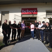 A imagem mostra um grupo alinhado em frente a um pr&eacute;dio de fachada clara com o letreiro &ldquo;bebec&ecirc;&rdquo; acima da entrada. As pessoas est&atilde;o posicionadas dos dois lados de uma faixa vermelha estendida, em um gesto simb&oacute;lico de inaugura&ccedil;&atilde;o. Grades met&aacute;licas, janelas com prote&ccedil;&atilde;o e parte externa pavimentada comp&otilde;em o cen&aacute;rio.