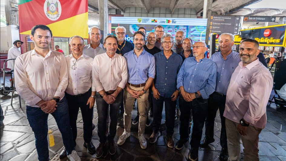 Grupo de cerca de 15 homens posando para foto dentro da Esta&ccedil;&atilde;o Rodovi&aacute;ria de Porto Alegre. Ao fundo, a bandeira do Rio Grande do Sul e um painel com logotipos institucionais do governo estadual. O ambiente &eacute; coberto, com estrutura de concreto aparente. As placas indicam &aacute;reas como "Embarque interestadual e internacional" e "Sala de Embarque".