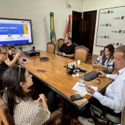 A imagem mostra uma reuni&atilde;o de trabalho em uma sala de reuni&otilde;es de estilo cl&aacute;ssico. Sete pessoas est&atilde;o sentadas ao redor de uma grande mesa de madeira retangular, participando de uma apresenta&ccedil;&atilde;o institucional. Um homem em destaque &agrave; direita fala enquanto opera um computador.

Detalhes dos Participantes
O Palestrante: No canto inferior direito, um homem de pele clara e cabelos curtos grisalhos, vestindo uma camisa social azul clara, gesticula com a m&atilde;o esquerda enquanto a direita opera um mouse sobre a mesa.

O Grupo: Outras seis pessoas (quatro mulheres e dois homens) est&atilde;o distribu&iacute;das ao redor da mesa. Algumas observam o palestrante, enquanto outras olham para uma tela ou para seus pr&oacute;prios celulares. Eles vestem roupas casuais e profissionais.

Mobili&aacute;rio: A mesa &eacute; de madeira clara com bordas escuras. As cadeiras s&atilde;o de madeira trabalhada com estofado em tom de couro marrom.

Elementos de Apoio e Ambiente
Monitor Digital: No fundo, &agrave; esquerda, h&aacute; um grande monitor de tela plana montado em um suporte m&oacute;vel. A tela exibe um slide com o t&iacute;tulo "DESTAQUES DA GEST&Atilde;O" e tr&ecirc;s pontos numerados sobre um fundo azul e branco.

Decora&ccedil;&atilde;o e S&iacute;mbolos: Ao fundo, no centro, est&atilde;o posicionadas duas bandeiras em mastros: a bandeira do Brasil e a bandeira do Rio Grande do Sul. &Agrave; direita, h&aacute; um grande painel branco (backdrop) com logotipos repetidos da Secretaria da Agricultura, Pecu&aacute;ria, Produ&ccedil;&atilde;o Sustent&aacute;vel e Irriga&ccedil;&atilde;o do Governo do Estado do Rio Grande do Sul.

Arquitetura: O piso &eacute; de madeira com um padr&atilde;o geom&eacute;trico de marchetaria (parquet). As paredes s&atilde;o brancas e h&aacute; uma porta de madeira escura ao fundo. Sobre a mesa, veem-se celulares, um telefone de confer&ecirc;ncia, um teclado preto, pap&eacute;is e copos de &aacute;gua.

Composi&ccedil;&atilde;o
A foto &eacute; tirada de um &acirc;ngulo superior lateral, capturando quase toda a extens&atilde;o da mesa e criando uma perspectiva que integra todos os participantes e os elementos institucionais da sala.