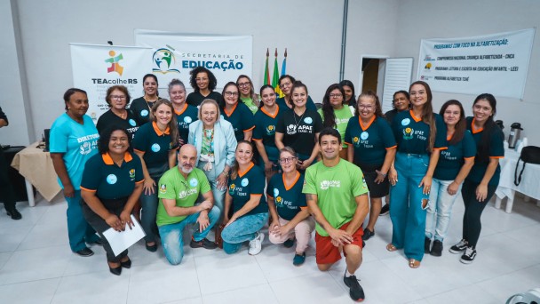 Esta &eacute; uma descri&ccedil;&atilde;o detalhada da imagem para fins de acessibilidade:

Vis&atilde;o Geral
A imagem mostra um grupo de aproximadamente 25 pessoas reunidas em um ambiente interno para uma fotografia coletiva. O grupo &eacute; composto majoritariamente por mulheres, muitas vestindo uniformes de uma institui&ccedil;&atilde;o. No centro, destaca-se uma autoridade governamental.

Detalhes do Grupo
Vestimentas: A maioria dos presentes usa camisetas de cor azul-petr&oacute;leo com golas laranjas ou camisetas verdes. Muitas dessas pe&ccedil;as exibem o logotipo da APAE Tapes.

Figura Central: No centro do grupo, est&aacute; uma mulher de pele clara, cabelos loiros curtos e &oacute;culos, vestindo um conjunto de blazer e cal&ccedil;a em tom azul-celeste claro. Ela sorri para a c&acirc;mera.

Disposi&ccedil;&atilde;o: As pessoas est&atilde;o organizadas em tr&ecirc;s n&iacute;veis: algumas ajoelhadas &agrave; frente, outras em p&eacute; no meio e uma fileira ao fundo. O clima &eacute; de celebra&ccedil;&atilde;o e uni&atilde;o.

Banners e Identifica&ccedil;&atilde;o
Ao fundo, tr&ecirc;s banners brancos fornecem o contexto do evento:

Banner &agrave; esquerda: Cont&eacute;m o logotipo colorido do programa TEAcolhe RS (Programa de Atendimento a Pessoas com Autismo).

Banner central: Exibe o logotipo da Secretaria de Educa&ccedil;&atilde;o do Governo do Estado do Rio Grande do Sul.

Banner &agrave; direita: Lista "Programas com foco na alfabetiza&ccedil;&atilde;o", mencionando iniciativas como o Compromisso Nacional Crian&ccedil;a Alfabetizada e o Programa Alfabetiza Tch&ecirc;.

Ambiente
Cen&aacute;rio: O local tem paredes brancas e ch&atilde;o de cer&acirc;mica clara. Ao fundo, entre os banners, s&atilde;o vis&iacute;veis as bandeiras do Brasil e do Rio Grande do Sul em mastros.

Composi&ccedil;&atilde;o: A ilumina&ccedil;&atilde;o &eacute; uniforme e direta. No canto inferior direito, &eacute; poss&iacute;vel ver parte de algumas bolsas e cadeiras, sugerindo que o grupo est&aacute; em um audit&oacute;rio ou sala de eventos multifuncional.