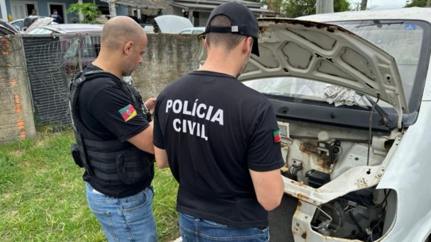 Foto mostra dois policiais civis revistando uma carca&ccedil;a de um autom&oacute;vel branco. Na camiseta preta de um dos policiais est&aacute; escrito "Pol&iacute;cia Civil".