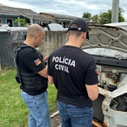 Foto mostra dois policiais civis revistando uma carca&ccedil;a de um autom&oacute;vel branco. Na camiseta preta de um dos policiais est&aacute; escrito "Pol&iacute;cia Civil".