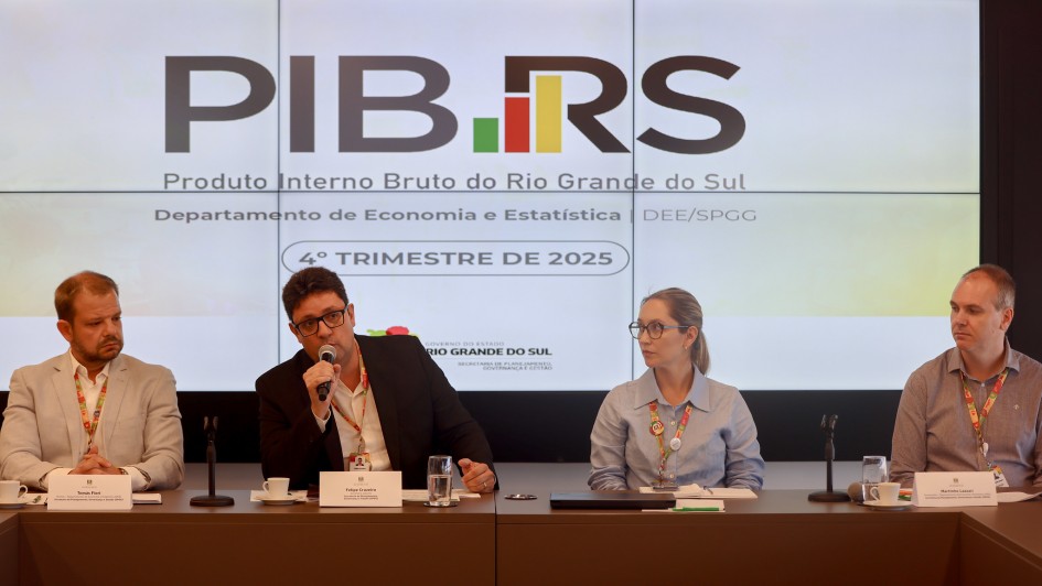 Esta &eacute; uma descri&ccedil;&atilde;o detalhada da imagem para fins de acessibilidade:

Vis&atilde;o Geral
A imagem mostra quatro integrantes do Governo do Estado do Rio Grande do Sul sentados em uma mesa de confer&ecirc;ncia durante uma coletiva de imprensa. Ao fundo, um painel digital apresenta dados sobre o crescimento econ&ocirc;mico do Estado.

Detalhes dos Participantes
Composi&ccedil;&atilde;o: Tr&ecirc;s homens e uma mulher est&atilde;o sentados lado a lado. Da esquerda para a direita:

Um homem de palet&oacute; claro e camisa branca.

Um homem de palet&oacute; escuro que fala ao microfone.

Uma mulher de camisa azul clara e &oacute;culos.

Um homem de camisa social cinza com padronagem mi&uacute;da.

Acess&oacute;rios: Todos utilizam crach&aacute;s com cord&otilde;es coloridos (verde, vermelho e amarelo, as cores da bandeira do RS). Sobre a mesa, h&aacute; placas de identifica&ccedil;&atilde;o com nomes, x&iacute;caras de caf&eacute;, copos de &aacute;gua e um notebook fechado.

Painel de Fundo
O tel&atilde;o central exibe informa&ccedil;&otilde;es do Departamento de Economia e Estat&iacute;stica (DEE/SPGG). Os elementos principais s&atilde;o:

T&iacute;tulo: Em letras grandes e pretas, l&ecirc;-se "PIB.RS". O ponto entre as palavras &eacute; substitu&iacute;do por um pequeno gr&aacute;fico de barras nas cores verde, vermelho e amarelo.

Subt&iacute;tulo: Logo abaixo, est&aacute; escrito "Produto Interno Bruto do Rio Grande do Sul".

Refer&ecirc;ncia Temporal: Um selo centralizado indica "4&ordm; TRIMESTRE DE 2025".

Logotipos: Na parte inferior do tel&atilde;o, aparece a marca oficial do Governo do Estado do Rio Grande do Sul.

Ambiente e Composi&ccedil;&atilde;o
Cen&aacute;rio: O ambiente &eacute; formal e bem iluminado. A mesa de reuni&otilde;es tem um acabamento em madeira escura.

Enquadramento: A foto &eacute; frontal e captura os participantes da cintura para cima, mantendo o foco n&iacute;tido tanto nas pessoas quanto nas informa&ccedil;&otilde;es exibidas no tel&atilde;o.