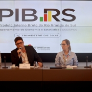 Esta &eacute; uma descri&ccedil;&atilde;o detalhada da imagem para fins de acessibilidade:

Vis&atilde;o Geral
A imagem mostra quatro integrantes do Governo do Estado do Rio Grande do Sul sentados em uma mesa de confer&ecirc;ncia durante uma coletiva de imprensa. Ao fundo, um painel digital apresenta dados sobre o crescimento econ&ocirc;mico do Estado.

Detalhes dos Participantes
Composi&ccedil;&atilde;o: Tr&ecirc;s homens e uma mulher est&atilde;o sentados lado a lado. Da esquerda para a direita:

Um homem de palet&oacute; claro e camisa branca.

Um homem de palet&oacute; escuro que fala ao microfone.

Uma mulher de camisa azul clara e &oacute;culos.

Um homem de camisa social cinza com padronagem mi&uacute;da.

Acess&oacute;rios: Todos utilizam crach&aacute;s com cord&otilde;es coloridos (verde, vermelho e amarelo, as cores da bandeira do RS). Sobre a mesa, h&aacute; placas de identifica&ccedil;&atilde;o com nomes, x&iacute;caras de caf&eacute;, copos de &aacute;gua e um notebook fechado.

Painel de Fundo
O tel&atilde;o central exibe informa&ccedil;&otilde;es do Departamento de Economia e Estat&iacute;stica (DEE/SPGG). Os elementos principais s&atilde;o:

T&iacute;tulo: Em letras grandes e pretas, l&ecirc;-se "PIB.RS". O ponto entre as palavras &eacute; substitu&iacute;do por um pequeno gr&aacute;fico de barras nas cores verde, vermelho e amarelo.

Subt&iacute;tulo: Logo abaixo, est&aacute; escrito "Produto Interno Bruto do Rio Grande do Sul".

Refer&ecirc;ncia Temporal: Um selo centralizado indica "4&ordm; TRIMESTRE DE 2025".

Logotipos: Na parte inferior do tel&atilde;o, aparece a marca oficial do Governo do Estado do Rio Grande do Sul.

Ambiente e Composi&ccedil;&atilde;o
Cen&aacute;rio: O ambiente &eacute; formal e bem iluminado. A mesa de reuni&otilde;es tem um acabamento em madeira escura.

Enquadramento: A foto &eacute; frontal e captura os participantes da cintura para cima, mantendo o foco n&iacute;tido tanto nas pessoas quanto nas informa&ccedil;&otilde;es exibidas no tel&atilde;o.