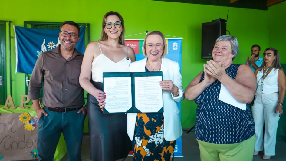 A imagem mostra um grupo reunido em frente a uma parede verde. No centro, duas pessoas seguram um documento aberto voltado para a c&acirc;mera. Ao fundo, h&aacute; banners institucionais e elementos de decora&ccedil;&atilde;o simples.