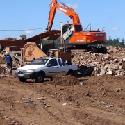 Processo de britagem de entulho, com escavadeira e m&aacute;quina de peneiramento operando sobre um grande volume de material demolido. Um ve&iacute;culo utilit&aacute;rio est&aacute; parado pr&oacute;ximo ao equipamento, e trabalhadores movimentam entulhos em meio ao solo revolvido.