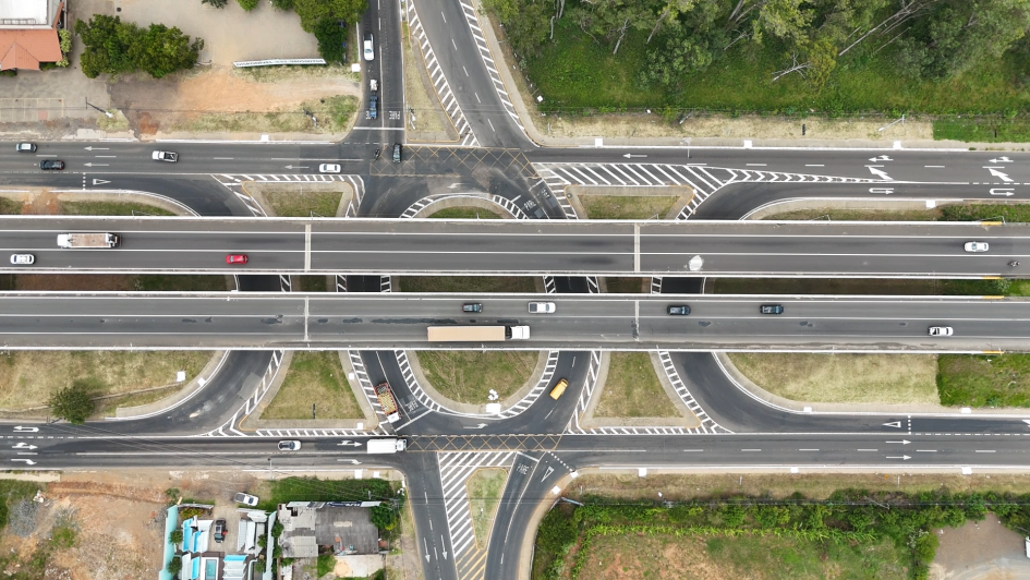 Esta &eacute; uma descri&ccedil;&atilde;o detalhada da imagem para fins de acessibilidade:

Vis&atilde;o Geral
A imagem &eacute; uma fotografia a&eacute;rea capturada diretamente de cima (vis&atilde;o vertical), mostrando um complexo entroncamento rodovi&aacute;rio na ERS-118. A composi&ccedil;&atilde;o &eacute; dominada por linhas asf&aacute;lticas cinzas, sinaliza&ccedil;&otilde;es brancas e &aacute;reas verdes de vegeta&ccedil;&atilde;o lateral.

Infraestrutura e Tra&ccedil;ado
Viadutos Centrais: Duas grandes pistas paralelas cruzam horizontalmente o centro da imagem. Elas s&atilde;o elevadas, funcionando como viadutos que permitem o fluxo cont&iacute;nuo de ve&iacute;culos.

Al&ccedil;as de Acesso: Abaixo dos viadutos, h&aacute; um sistema de al&ccedil;as de retorno e acesso em formato circular e curvo, permitindo que os ve&iacute;culos troquem de dire&ccedil;&atilde;o ou acessem as vias laterais.

Sinaliza&ccedil;&atilde;o: As pistas apresentam sinaliza&ccedil;&otilde;es horizontais n&iacute;tidas, incluindo faixas brancas cont&iacute;nuas e pontilhadas, setas de dire&ccedil;&atilde;o, faixas de pedestres e &aacute;reas zebradas em amarelo (canaliza&ccedil;&atilde;o de tr&aacute;fego) nos pontos de bifurca&ccedil;&atilde;o.

Tr&aacute;fego e Ve&iacute;culos
V&aacute;rios ve&iacute;culos est&atilde;o em movimento ou posicionados nas vias, ajudando a dar escala &agrave; infraestrutura:

Um caminh&atilde;o de carga branco com ba&uacute; longo transita na pista central inferior.

Carros de passeio de diversas cores (branco, vermelho, prata e preto) est&atilde;o espalhados pelas diferentes al&ccedil;as e pistas principais.

Um caminh&atilde;o menor transportando o que parece ser carga viva ou sacos coloridos &eacute; vis&iacute;vel em uma das curvas de acesso.

Elementos do Entorno
Vegeta&ccedil;&atilde;o: No canto superior direito e inferior direito, h&aacute; &aacute;reas de mata densa com &aacute;rvores verdes. &Aacute;reas de gramado rasteiro preenchem os v&atilde;os entre as al&ccedil;as de acesso.

&Aacute;rea Urbana: No canto superior esquerdo e inferior esquerdo, observam-se telhados de edifica&ccedil;&otilde;es comerciais ou industriais, p&aacute;tios de estacionamento e solo exposto em tons de marrom e bege, sugerindo &aacute;reas em obras ou dep&oacute;sitos de materiais.

Composi&ccedil;&atilde;o e Ilumina&ccedil;&atilde;o
A luz &eacute; zenital (meio-dia), o que minimiza as sombras projetadas pelos ve&iacute;culos e viadutos, proporcionando uma vis&atilde;o clara e t&eacute;cnica de toda a engenharia do local. A simetria das pistas centrais cria um forte eixo horizontal na imagem.