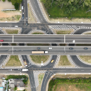 Esta &eacute; uma descri&ccedil;&atilde;o detalhada da imagem para fins de acessibilidade:

Vis&atilde;o Geral
A imagem &eacute; uma fotografia a&eacute;rea capturada diretamente de cima (vis&atilde;o vertical), mostrando um complexo entroncamento rodovi&aacute;rio na ERS-118. A composi&ccedil;&atilde;o &eacute; dominada por linhas asf&aacute;lticas cinzas, sinaliza&ccedil;&otilde;es brancas e &aacute;reas verdes de vegeta&ccedil;&atilde;o lateral.

Infraestrutura e Tra&ccedil;ado
Viadutos Centrais: Duas grandes pistas paralelas cruzam horizontalmente o centro da imagem. Elas s&atilde;o elevadas, funcionando como viadutos que permitem o fluxo cont&iacute;nuo de ve&iacute;culos.

Al&ccedil;as de Acesso: Abaixo dos viadutos, h&aacute; um sistema de al&ccedil;as de retorno e acesso em formato circular e curvo, permitindo que os ve&iacute;culos troquem de dire&ccedil;&atilde;o ou acessem as vias laterais.

Sinaliza&ccedil;&atilde;o: As pistas apresentam sinaliza&ccedil;&otilde;es horizontais n&iacute;tidas, incluindo faixas brancas cont&iacute;nuas e pontilhadas, setas de dire&ccedil;&atilde;o, faixas de pedestres e &aacute;reas zebradas em amarelo (canaliza&ccedil;&atilde;o de tr&aacute;fego) nos pontos de bifurca&ccedil;&atilde;o.

Tr&aacute;fego e Ve&iacute;culos
V&aacute;rios ve&iacute;culos est&atilde;o em movimento ou posicionados nas vias, ajudando a dar escala &agrave; infraestrutura:

Um caminh&atilde;o de carga branco com ba&uacute; longo transita na pista central inferior.

Carros de passeio de diversas cores (branco, vermelho, prata e preto) est&atilde;o espalhados pelas diferentes al&ccedil;as e pistas principais.

Um caminh&atilde;o menor transportando o que parece ser carga viva ou sacos coloridos &eacute; vis&iacute;vel em uma das curvas de acesso.

Elementos do Entorno
Vegeta&ccedil;&atilde;o: No canto superior direito e inferior direito, h&aacute; &aacute;reas de mata densa com &aacute;rvores verdes. &Aacute;reas de gramado rasteiro preenchem os v&atilde;os entre as al&ccedil;as de acesso.

&Aacute;rea Urbana: No canto superior esquerdo e inferior esquerdo, observam-se telhados de edifica&ccedil;&otilde;es comerciais ou industriais, p&aacute;tios de estacionamento e solo exposto em tons de marrom e bege, sugerindo &aacute;reas em obras ou dep&oacute;sitos de materiais.

Composi&ccedil;&atilde;o e Ilumina&ccedil;&atilde;o
A luz &eacute; zenital (meio-dia), o que minimiza as sombras projetadas pelos ve&iacute;culos e viadutos, proporcionando uma vis&atilde;o clara e t&eacute;cnica de toda a engenharia do local. A simetria das pistas centrais cria um forte eixo horizontal na imagem.