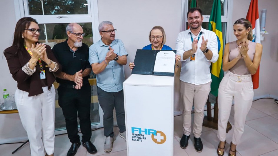Imagem mostra seis pessoas de p&eacute; lado a lado em uma sala, com cinco delas batendo palmas e uma, mais ao centro, mostrando um documento.