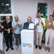 Imagem mostra seis pessoas de p&eacute; lado a lado em uma sala, com cinco delas batendo palmas e uma, mais ao centro, mostrando um documento.