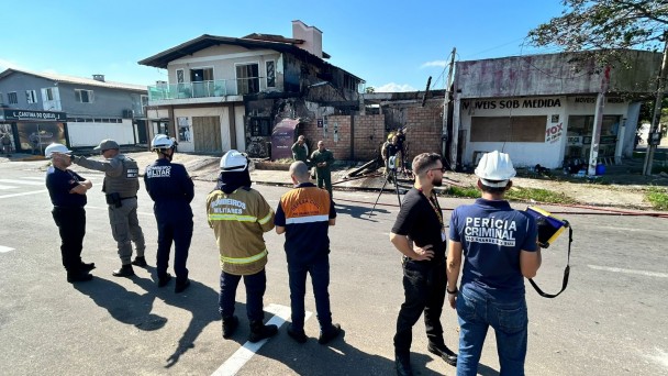 Um grupo de sete pessoas sob uma lona observa uma casa com avarias em sua fachada