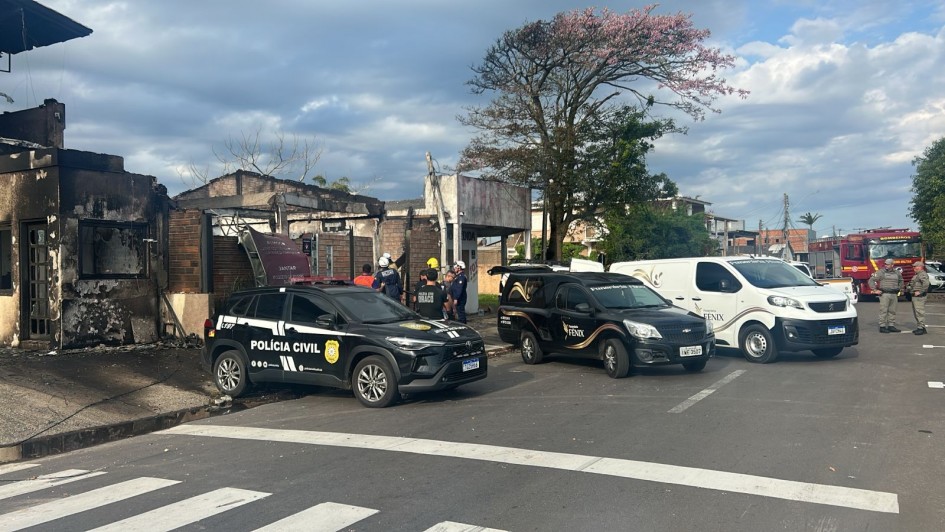 Tr&ecirc;s ve&iacute;culos estacionados de r&eacute; junto &agrave; cal&ccedil;ada de uma casa parcialmente destru&iacute;da. No primeiro plano, uma caminhonete da Pol&iacute;cia Civil, ao lado dois ve&iacute;culos funer&aacute;rios, mais ao fundo caminh&otilde;es dos bombeiros. Junto aos ve&iacute;culos e na rua, v&aacute;rios policiais e bombeiros 