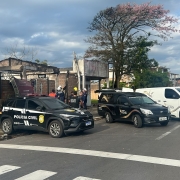 Tr&ecirc;s ve&iacute;culos estacionados de r&eacute; junto &agrave; cal&ccedil;ada de uma casa parcialmente destru&iacute;da. No primeiro plano, uma caminhonete da Pol&iacute;cia Civil, ao lado dois ve&iacute;culos funer&aacute;rios, mais ao fundo caminh&otilde;es dos bombeiros. Junto aos ve&iacute;culos e na rua, v&aacute;rios policiais e bombeiros 