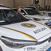 Viaturas da Brigada Militar alinhadas em &aacute;rea coberta, com padroniza&ccedil;&atilde;o visual branca, faixas amarelas e identifica&ccedil;&atilde;o policial. Os ve&iacute;culos est&atilde;o estacionados lado a lado, com ilumina&ccedil;&atilde;o interna do pavilh&atilde;o e estrutura administrativa ao fundo.