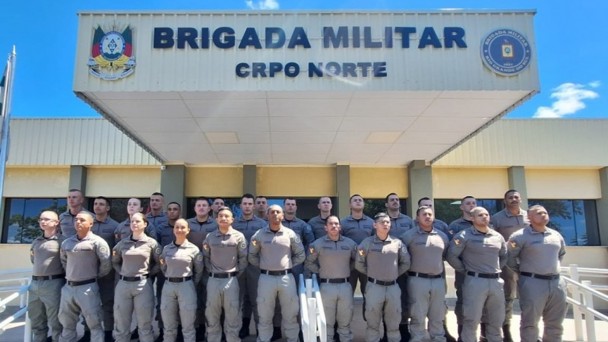 A fotografia colorida mostra um grupo de policiais militares em frente a uma sede da corpora&ccedil;&atilde;o.

Detalhes da cena:

O Grupo: Cerca de 25 policiais (homens e mulheres) est&atilde;o posicionados em forma&ccedil;&atilde;o para uma foto oficial. Eles est&atilde;o organizados em tr&ecirc;s fileiras horizontais, em degraus, todos em p&eacute;, com as m&atilde;os para tr&aacute;s e express&atilde;o s&eacute;ria.

O Uniforme: Todos vestem o uniforme operacional da Brigada Militar do Rio Grande do Sul, composto por camisas e cal&ccedil;as em tom cinza-bandeirante, cintos pretos e botas pretas.

O Pr&eacute;dio: Ao fundo, o grupo est&aacute; posicionado sob a marquise de um pr&eacute;dio de cor clara. Na fachada da marquise, l&ecirc;-se em letras pretas e grandes: "BRIGADA MILITAR", e logo abaixo, em letras menores, "CRPO NORTE". O bras&atilde;o oficial da corpora&ccedil;&atilde;o est&aacute; vis&iacute;vel &agrave; esquerda e &agrave; direita do letreiro.

Ambiente e Ilumina&ccedil;&atilde;o: A foto foi tirada sob luz solar intensa e c&eacute;u azul com poucas nuvens. As sombras est&atilde;o projetadas diretamente abaixo dos policiais, indicando que o sol est&aacute; em uma posi&ccedil;&atilde;o elevada. H&aacute; rampas de acessibilidade com corrim&atilde;os brancos nas laterais do grupo.