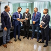 Imagem mostra cinco homens em p&eacute; em uma sala de espera com dois deles trocando documentos.