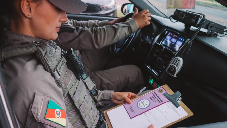 Foto mostra policial militar sentada na carona de uma viatura. Ela est&aacute; com uma prancheta com um panfelto lil&aacute;s. Outro policial est&aacute; sentado ao volante. Dentro do carro, h&aacute; um r&aacute;dio comunicador. 