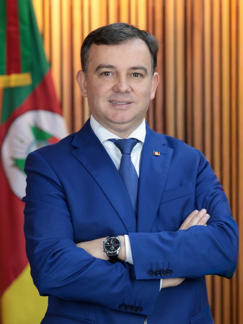 Homem de gravata e palet&oacute; azul pousa para foto oficial, &agrave; frente de uma bandeira do Rio Grande do Sul