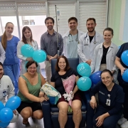A fotografia colorida registra um momento de celebra&ccedil;&atilde;o em um ambiente hospitalar. Um grupo de doze pessoas rodeia uma mulher sentada ao centro, que sorri enquanto segura um buqu&ecirc; de flores.

Detalhes da cena:

A paciente: Uma mulher de cabelos castanhos claros est&aacute; sentada em uma poltrona hospitalar azul. Ela segura um buqu&ecirc; de flores envolto em papel rosa e uma sacola de presente.

A equipe de sa&uacute;de e acompanhantes: Ao redor dela, dez profissionais de sa&uacute;de e um homem em trajes civis posam para a foto. Os profissionais vestem jalecos brancos com estetosc&oacute;pios no pesco&ccedil;o ou uniformes privativos (scrubs) em tons de azul claro e azul marinho. Todos sorriem para a c&acirc;mera.

Decora&ccedil;&atilde;o festiva: O grupo segura diversos bal&otilde;es azuis inflados. Alguns bal&otilde;es possuem o logotipo "HCI - Hospital de Cl&iacute;nicas Iju&iacute;" impresso em branco.

O ambiente: A cena ocorre dentro de um quarto de hospital. Ao fundo, v&ecirc;-se uma janela com persianas horizontais fechadas e um suporte de soro com uma bolsa de fluido pendurada, conectada &agrave; paciente.

Ilumina&ccedil;&atilde;o: A luz &eacute; clara e uniforme, caracter&iacute;stica de ambientes cl&iacute;nicos, transmitindo uma atmosfera de al&iacute;vio e comemora&ccedil;&atilde;o.