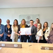 A fotografia colorida mostra um grupo de nove pessoas (tr&ecirc;s homens e seis mulheres) em p&eacute;, posicionadas lado a lado em um ambiente de escrit&oacute;rio, celebrando a assinatura de um acordo.

Detalhes da cena:

O ato central: No centro do grupo, uma mulher loira vestindo um blazer estampado e um homem de &oacute;culos vestindo um palet&oacute; cinza seguram juntos um documento oficial aberto. Eles sorriem para a c&acirc;mera, simbolizando a formaliza&ccedil;&atilde;o de uma parceria. O documento exibe o bras&atilde;o do Estado do Rio Grande do Sul no topo.

Os participantes: As outras sete pessoas est&atilde;o distribu&iacute;das ao redor do casal central. Elas vestem trajes que variam entre o casual e o formal (blazers, camisas sociais e blusas coloridas). V&aacute;rias pessoas no grupo utilizam crach&aacute;s com cord&otilde;es vermelhos e amarelos pendurados no pesco&ccedil;o.

O ambiente: O grupo est&aacute; atr&aacute;s de uma bancada de madeira clara com tomadas embutidas. Ao fundo, em uma parede branca, h&aacute; uma placa de acr&iacute;lico transparente com um bras&atilde;o circular e o texto: "CONSELHO REGIONAL DE FISIOTERAPIA E TERAPIA OCUPACIONAL DO RIO GRANDE DO SUL".

Ilumina&ccedil;&atilde;o: A foto possui ilumina&ccedil;&atilde;o frontal clara e sombras suaves, com um clima profissional e positivo. Uma caneta preta est&aacute; apoiada sobre a bancada de madeira, em frente ao grupo.