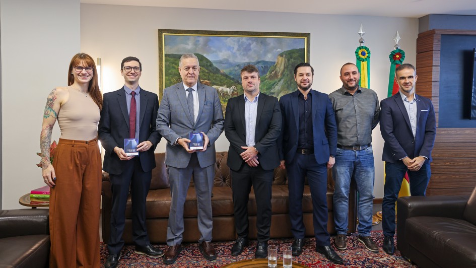 A fotografia colorida mostra um grupo de sete pessoas (uma mulher e seis homens) em p&eacute;, lado a lado, em um ambiente de escrit&oacute;rio formal ou gabinete. Eles est&atilde;o posicionados para uma foto oficial de entrega de documento.

Detalhes da cena:

As pessoas e o ato: O grupo est&aacute; organizado horizontalmente. Dois homens, posicionados &agrave; esquerda do centro, seguram pequenos livretos ou documentos de capa azul e detalhes brancos. A mulher, &agrave; extrema esquerda, tem cabelos ruivos e tatuagens vis&iacute;veis no bra&ccedil;o direito, vestindo uma regata bege e cal&ccedil;a pantalona marrom. Os homens vestem predominantemente trajes formais, como ternos, palet&oacute;s e camisas sociais, com exce&ccedil;&atilde;o de um homem &agrave; direita que veste camisa cinza e cal&ccedil;a jeans.

O ambiente: Ao fundo, no centro, h&aacute; uma grande pintura emoldurada representando uma paisagem de campo com gado e montanhas. &Agrave; direita, est&atilde;o posicionadas duas bandeiras oficiais em mastros (Brasil e Rio Grande do Sul). O mobili&aacute;rio inclui um sof&aacute; de couro marrom atr&aacute;s do grupo, poltronas de couro e uma mesa de centro de madeira em primeiro plano, onde se veem dois copos com &aacute;gua.

Ch&atilde;o e Paredes: O grupo est&aacute; sobre um tapete cl&aacute;ssico com padr&otilde;es detalhados. As paredes s&atilde;o de tom neutro claro, e h&aacute; detalhes em madeira escura nas colunas &agrave; direita.

Ilumina&ccedil;&atilde;o: A luz &eacute; interna e bem distribu&iacute;da, real&ccedil;ando as express&otilde;es serenas e profissionais de todos os presentes. No canto esquerdo, h&aacute; uma lumin&aacute;ria de coluna acesa.