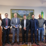 A fotografia colorida mostra um grupo de sete pessoas (uma mulher e seis homens) em p&eacute;, lado a lado, em um ambiente de escrit&oacute;rio formal ou gabinete. Eles est&atilde;o posicionados para uma foto oficial de entrega de documento.

Detalhes da cena:

As pessoas e o ato: O grupo est&aacute; organizado horizontalmente. Dois homens, posicionados &agrave; esquerda do centro, seguram pequenos livretos ou documentos de capa azul e detalhes brancos. A mulher, &agrave; extrema esquerda, tem cabelos ruivos e tatuagens vis&iacute;veis no bra&ccedil;o direito, vestindo uma regata bege e cal&ccedil;a pantalona marrom. Os homens vestem predominantemente trajes formais, como ternos, palet&oacute;s e camisas sociais, com exce&ccedil;&atilde;o de um homem &agrave; direita que veste camisa cinza e cal&ccedil;a jeans.

O ambiente: Ao fundo, no centro, h&aacute; uma grande pintura emoldurada representando uma paisagem de campo com gado e montanhas. &Agrave; direita, est&atilde;o posicionadas duas bandeiras oficiais em mastros (Brasil e Rio Grande do Sul). O mobili&aacute;rio inclui um sof&aacute; de couro marrom atr&aacute;s do grupo, poltronas de couro e uma mesa de centro de madeira em primeiro plano, onde se veem dois copos com &aacute;gua.

Ch&atilde;o e Paredes: O grupo est&aacute; sobre um tapete cl&aacute;ssico com padr&otilde;es detalhados. As paredes s&atilde;o de tom neutro claro, e h&aacute; detalhes em madeira escura nas colunas &agrave; direita.

Ilumina&ccedil;&atilde;o: A luz &eacute; interna e bem distribu&iacute;da, real&ccedil;ando as express&otilde;es serenas e profissionais de todos os presentes. No canto esquerdo, h&aacute; uma lumin&aacute;ria de coluna acesa.