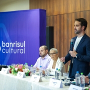 A fotografia colorida mostra o governador Eduardo Leite discursando durante um evento institucional. Ele est&aacute; em p&eacute;, posicionado &agrave; direita do centro da imagem, falando ao microfone.

Detalhes da cena:

O orador: Eduardo Leite tem barba curta, cabelos escuros e veste um palet&oacute; azul-marinho sobre uma camiseta branca. Ele segura um microfone com a m&atilde;o direita e gesticula com a m&atilde;o esquerda enquanto olha para a plateia (fora do campo de vis&atilde;o).

A mesa de honra: &Agrave; frente do governador, h&aacute; uma mesa comprida coberta com uma toalha branca rendada. Sentados &agrave; mesa, observando o discurso, est&atilde;o outras tr&ecirc;s pessoas: um homem de camisa lil&aacute;s, uma mulher de &oacute;culos e blazer branco, e um homem de terno cinza e gravata azul. Sobre a mesa, h&aacute; arranjos de flores coloridas, copos de cristal, garrafas de &aacute;gua e placas de identifica&ccedil;&atilde;o.

O cen&aacute;rio: Ao fundo, &agrave; esquerda, destaca-se um painel vertical azul vibrante com o logotipo "banrisul cultural" em branco. &Agrave; direita, a parede &eacute; revestida com pain&eacute;is de madeira de tom m&eacute;dio.

Ilumina&ccedil;&atilde;o e composi&ccedil;&atilde;o: A ilumina&ccedil;&atilde;o &eacute; direta e clara, t&iacute;pica de eventos em audit&oacute;rios ou centros de conven&ccedil;&otilde;es. A foto captura o governador da cintura para cima, com o p&uacute;blico e os demais integrantes da mesa compondo o contexto da cerim&ocirc;nia.