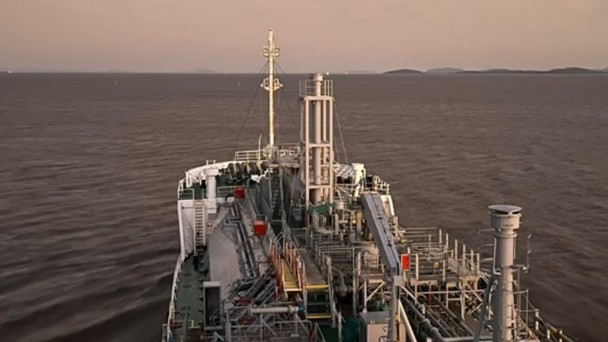 A fotografia colorida mostra a perspectiva da proa de um grande navio cargueiro navegando em &aacute;guas calmas.

Detalhes da cena:

O Navio: A imagem &eacute; capturada de um ponto elevado da embarca&ccedil;&atilde;o, olhando para a frente. O conv&eacute;s &eacute; repleto de estruturas met&aacute;licas complexas, incluindo tubula&ccedil;&otilde;es, passarelas de gradeado, escadas e colunas verticais pintadas de cinza e branco. H&aacute; detalhes em vermelho em algumas caixas e v&aacute;lvulas.

A &Aacute;gua: O navio navega por uma vasta extens&atilde;o de &aacute;gua escura, que reflete as cores suaves do c&eacute;u. Marcas de espuma branca s&atilde;o vis&iacute;veis nas laterais, indicando o movimento da embarca&ccedil;&atilde;o.

O C&eacute;u e o Horizonte: O c&eacute;u apresenta tons de bege e cinza, sugerindo o crep&uacute;sculo ou o amanhecer. No horizonte distante, &agrave; direita, &eacute; poss&iacute;vel ver silhuetas baixas de terra firme ou ilhas sob uma luz difusa.

Composi&ccedil;&atilde;o e Ilumina&ccedil;&atilde;o: A ilumina&ccedil;&atilde;o &eacute; suave e indireta, vinda do c&eacute;u nublado, o que minimiza sombras fortes e destaca a silhueta industrial do navio contra a natureza ao redor. O &acirc;ngulo da foto cria uma sensa&ccedil;&atilde;o de dire&ccedil;&atilde;o e continuidade da jornada.