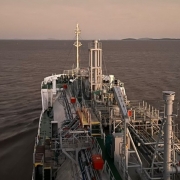 A fotografia colorida mostra a perspectiva da proa de um grande navio cargueiro navegando em &aacute;guas calmas.

Detalhes da cena:

O Navio: A imagem &eacute; capturada de um ponto elevado da embarca&ccedil;&atilde;o, olhando para a frente. O conv&eacute;s &eacute; repleto de estruturas met&aacute;licas complexas, incluindo tubula&ccedil;&otilde;es, passarelas de gradeado, escadas e colunas verticais pintadas de cinza e branco. H&aacute; detalhes em vermelho em algumas caixas e v&aacute;lvulas.

A &Aacute;gua: O navio navega por uma vasta extens&atilde;o de &aacute;gua escura, que reflete as cores suaves do c&eacute;u. Marcas de espuma branca s&atilde;o vis&iacute;veis nas laterais, indicando o movimento da embarca&ccedil;&atilde;o.

O C&eacute;u e o Horizonte: O c&eacute;u apresenta tons de bege e cinza, sugerindo o crep&uacute;sculo ou o amanhecer. No horizonte distante, &agrave; direita, &eacute; poss&iacute;vel ver silhuetas baixas de terra firme ou ilhas sob uma luz difusa.

Composi&ccedil;&atilde;o e Ilumina&ccedil;&atilde;o: A ilumina&ccedil;&atilde;o &eacute; suave e indireta, vinda do c&eacute;u nublado, o que minimiza sombras fortes e destaca a silhueta industrial do navio contra a natureza ao redor. O &acirc;ngulo da foto cria uma sensa&ccedil;&atilde;o de dire&ccedil;&atilde;o e continuidade da jornada.