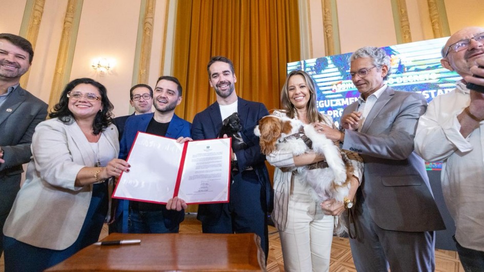 Foto mostra o governador Eduardo Leite segurando um pequeno cachorro preto e o vice Gabriel Leite, junto com a secret&aacute;ria que seguram um documento assinado. Outra mulher segura um cachorro branco e manchas marrons. Outras autoridades est&atilde;o juntos e aplaudem a cena. 