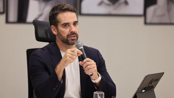 Esta &eacute; uma descri&ccedil;&atilde;o da imagem com foco em acessibilidade:

A fotografia colorida mostra um plano m&eacute;dio do governador Eduardo Leite durante uma atividade oficial de trabalho.

Detalhes da cena:

O orador: Eduardo Leite, um homem branco com barba e cabelos castanhos curtos, est&aacute; sentado &agrave; mesa. Ele veste um palet&oacute; azul-marinho sobre uma camiseta branca. Com a m&atilde;o esquerda, ele segura um microfone prateado pr&oacute;ximo &agrave; boca, enquanto a m&atilde;o direita gesticula, segurando uma caneta branca, indicando que est&aacute; em meio a uma explica&ccedil;&atilde;o. Ele usa um rel&oacute;gio de pulso com pulseira clara.

A mesa de trabalho: &Agrave; sua frente, sobre a mesa de tom claro, h&aacute; um tablet (iPad) apoiado em uma capa-teclado preta, pap&eacute;is de trabalho e um copo de vidro com &aacute;gua.

O ambiente: O fundo &eacute; composto por uma parede de cor neutra (cinza claro). Atr&aacute;s do governador, &eacute; poss&iacute;vel ver parte de fotografias em preto e branco emolduradas, que decoram o ambiente da reuni&atilde;o.

Composi&ccedil;&atilde;o e Ilumina&ccedil;&atilde;o: A imagem tem um enquadramento focado no governador, com o fundo levemente desfocado para dar destaque &agrave; sua express&atilde;o e fala. A