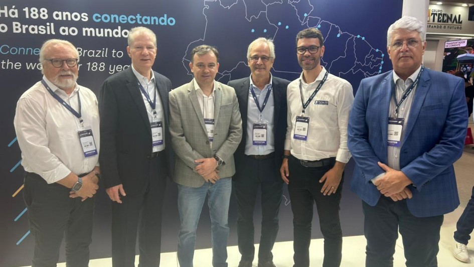 A fotografia colorida mostra um grupo de seis homens em p&eacute;, posicionados lado a lado, em frente a um painel institucional durante um evento de log&iacute;stica.

Detalhes da cena:

Os participantes: Os seis homens est&atilde;o alinhados horizontalmente e olham para a c&acirc;mera. Eles vestem trajes que variam entre o social e o casual de neg&oacute;cios (palet&oacute;s, blazers, camisas sociais e cal&ccedil;as jeans ou de sarja). Todos utilizam crach&aacute;s oficiais do evento pendurados no pesco&ccedil;o com cord&otilde;es azuis.

O painel de fundo: Atr&aacute;s do grupo, h&aacute; um grande painel azul escuro. No centro, destaca-se um mapa do Brasil desenhado em linhas brancas finas, com pontos azuis indicando localiza&ccedil;&otilde;es espec&iacute;ficas. &Agrave; esquerda do mapa, l&ecirc;-se a frase em branco e azul claro: "H&aacute; 188 anos conectando o Brasil ao mundo", com a tradu&ccedil;&atilde;o em ingl&ecirc;s logo abaixo.

O ambiente: O grupo est&aacute; em um pavilh&atilde;o de feira de neg&oacute;cios. No canto superior direito, &eacute; poss&iacute;vel ver parte de um estande vizinho com o logotipo "GRUPO TECNAL". O ch&atilde;o &eacute; claro e a ilumina&ccedil;&atilde;o &eacute; direta e intensa, t&iacute;pica de grandes centros de exposi&ccedil;&otilde;es.

Composi&ccedil;&atilde;o e Ilumina&ccedil;&atilde;o: A foto &eacute; frontal e captura o grupo da altura dos joelhos para cima. A luz reflete levemente no painel ao fundo e destaca a formalidade da agenda estrat&eacute;gica entre os participantes.