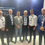 A fotografia colorida mostra um grupo de seis homens em p&eacute;, posicionados lado a lado, em frente a um painel institucional durante um evento de log&iacute;stica.

Detalhes da cena:

Os participantes: Os seis homens est&atilde;o alinhados horizontalmente e olham para a c&acirc;mera. Eles vestem trajes que variam entre o social e o casual de neg&oacute;cios (palet&oacute;s, blazers, camisas sociais e cal&ccedil;as jeans ou de sarja). Todos utilizam crach&aacute;s oficiais do evento pendurados no pesco&ccedil;o com cord&otilde;es azuis.

O painel de fundo: Atr&aacute;s do grupo, h&aacute; um grande painel azul escuro. No centro, destaca-se um mapa do Brasil desenhado em linhas brancas finas, com pontos azuis indicando localiza&ccedil;&otilde;es espec&iacute;ficas. &Agrave; esquerda do mapa, l&ecirc;-se a frase em branco e azul claro: "H&aacute; 188 anos conectando o Brasil ao mundo", com a tradu&ccedil;&atilde;o em ingl&ecirc;s logo abaixo.

O ambiente: O grupo est&aacute; em um pavilh&atilde;o de feira de neg&oacute;cios. No canto superior direito, &eacute; poss&iacute;vel ver parte de um estande vizinho com o logotipo "GRUPO TECNAL". O ch&atilde;o &eacute; claro e a ilumina&ccedil;&atilde;o &eacute; direta e intensa, t&iacute;pica de grandes centros de exposi&ccedil;&otilde;es.

Composi&ccedil;&atilde;o e Ilumina&ccedil;&atilde;o: A foto &eacute; frontal e captura o grupo da altura dos joelhos para cima. A luz reflete levemente no painel ao fundo e destaca a formalidade da agenda estrat&eacute;gica entre os participantes.