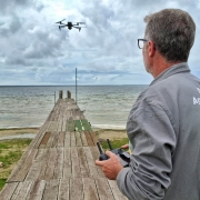 A fotografia colorida mostra um homem operando um drone em uma &aacute;rea natural de preserva&ccedil;&atilde;o, sob um c&eacute;u nublado.

Detalhes da cena:

O operador: Um homem de cabelos grisalhos e &oacute;culos &eacute; visto de costas e de perfil, do lado direito da imagem. Ele veste um macac&atilde;o cinza de trabalho com as palavras "Defesa Agropecu&aacute;ria" impressas em branco nas costas. Ele segura e opera um controle remoto com tela integrada.

O drone: Um drone quadric&oacute;ptero de cor escura est&aacute; voando no ar, posicionado na parte superior central da imagem, contra as nuvens cinzas e brancas.

O ambiente: A cena ocorre na Esta&ccedil;&atilde;o Ecol&oacute;gica do Taim. O homem est&aacute; em p&eacute; sobre um trapiche (pier) de madeira r&uacute;stica que se estende em dire&ccedil;&atilde;o a uma grande lagoa de &aacute;guas calmas. A vegeta&ccedil;&atilde;o nas margens &eacute; rasteira e verde, com algumas &aacute;reas de areia clara.

O horizonte: Ao fundo, a linha do horizonte divide a lagoa do c&eacute;u densamente nublado, transmitindo uma sensa&ccedil;&atilde;o de vastid&atilde;o e isolamento natural.

Ilumina&ccedil;&atilde;o: A luz &eacute; difusa e natural, t&iacute;pica de um dia encoberto, o que suaviza as cores da paisagem e destaca os elementos tecnol&oacute;gicos (drone e controle) em meio ao ambiente r&uacute;stico.