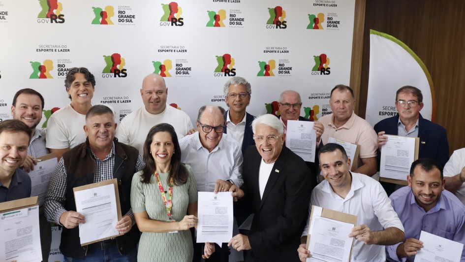 Esta &eacute; uma descri&ccedil;&atilde;o da imagem com foco em acessibilidade:

A fotografia colorida captura um momento de celebra&ccedil;&atilde;o coletiva ap&oacute;s a assinatura de atos oficiais entre o governo estadual e representantes municipais.

Detalhes da cena:

Os participantes e o ato: Um grupo numeroso de pessoas, majoritariamente homens, posa sorridente para a c&acirc;mera. Muitos deles seguram folhas de papel com o bras&atilde;o do Estado, representando os conv&ecirc;nios assinados. No centro, destaca-se uma mulher de vestido verde claro e um homem de cabelos brancos, &oacute;culos e terno preto, que seguram juntos um dos documentos.

O painel de fundo: Atr&aacute;s do grupo, h&aacute; um grande painel branco repetindo diversas vezes os logotipos oficiais: "GOV RS" (com silhuetas coloridas de rostos em vermelho, amarelo e verde) e "SECRETARIA DO ESPORTE E LAZER".

O ambiente: A cena ocorre em um ambiente interno com teto de placas brancas e ilumina&ccedil;&atilde;o embutida. &Agrave; direita, nota-se uma parede com pain&eacute;is de madeira e parte de um banner vertical.

Composi&ccedil;&atilde;o e ilumina&ccedil;&atilde;o: A foto &eacute; frontal e agrupa as pessoas de forma pr&oacute;xima para que todas apare&ccedil;am no enquadramento. A ilumina&ccedil;&atilde;o &eacute; clara e uniforme, t&iacute;pica de salas de reuni&otilde;es governamentais.
