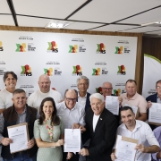 Esta &eacute; uma descri&ccedil;&atilde;o da imagem com foco em acessibilidade:

A fotografia colorida captura um momento de celebra&ccedil;&atilde;o coletiva ap&oacute;s a assinatura de atos oficiais entre o governo estadual e representantes municipais.

Detalhes da cena:

Os participantes e o ato: Um grupo numeroso de pessoas, majoritariamente homens, posa sorridente para a c&acirc;mera. Muitos deles seguram folhas de papel com o bras&atilde;o do Estado, representando os conv&ecirc;nios assinados. No centro, destaca-se uma mulher de vestido verde claro e um homem de cabelos brancos, &oacute;culos e terno preto, que seguram juntos um dos documentos.

O painel de fundo: Atr&aacute;s do grupo, h&aacute; um grande painel branco repetindo diversas vezes os logotipos oficiais: "GOV RS" (com silhuetas coloridas de rostos em vermelho, amarelo e verde) e "SECRETARIA DO ESPORTE E LAZER".

O ambiente: A cena ocorre em um ambiente interno com teto de placas brancas e ilumina&ccedil;&atilde;o embutida. &Agrave; direita, nota-se uma parede com pain&eacute;is de madeira e parte de um banner vertical.

Composi&ccedil;&atilde;o e ilumina&ccedil;&atilde;o: A foto &eacute; frontal e agrupa as pessoas de forma pr&oacute;xima para que todas apare&ccedil;am no enquadramento. A ilumina&ccedil;&atilde;o &eacute; clara e uniforme, t&iacute;pica de salas de reuni&otilde;es governamentais.