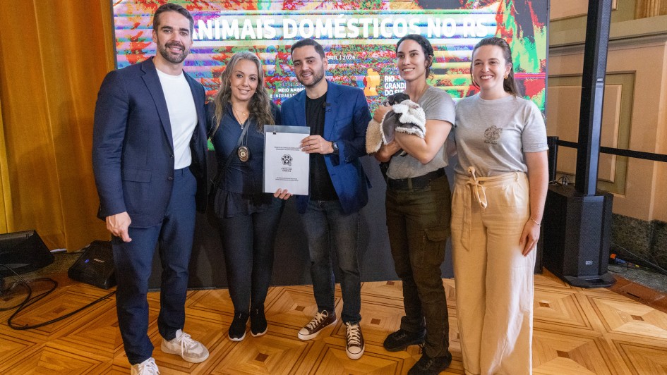 Esta &eacute; uma descri&ccedil;&atilde;o da imagem com foco em acessibilidade:

A fotografia colorida captura um momento de intera&ccedil;&atilde;o afetuosa entre duas mulheres e um cachorro de pequeno porte, em um ambiente interno iluminado.

Detalhes da cena:

As participantes: &Agrave; esquerda, uma mulher de cabelos loiros e &oacute;culos sorri enquanto observa o animal. &Agrave; direita, uma mulher de cabelos escuros segura o cachorro no colo, aproximando o rosto dele para um "beijo" ou carinho.

O animal: Um c&atilde;o vira-lata (SRD) de pelagem curta, predominantemente branca com manchas marrons na cabe&ccedil;a e nas orelhas. Ele est&aacute; calmo no colo, com as patas dianteiras apoiadas no bra&ccedil;o da mulher.

O ambiente: O fundo &eacute; composto por uma parede clara e, &agrave; direita, uma estrutura de madeira com o que parece ser um bras&atilde;o ou ins&iacute;gnia oficial fixada, sugerindo um gabinete ou &oacute;rg&atilde;o p&uacute;blico.

Composi&ccedil;&atilde;o e ilumina&ccedil;&atilde;o: A foto &eacute; um plano m&eacute;dio, focando da cintura para cima das participantes. A ilumina&ccedil;&atilde;o &eacute; suave e frontal, destacando as express&otilde;es de alegria e o cuidado com o animal.
