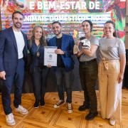 Esta &eacute; uma descri&ccedil;&atilde;o da imagem com foco em acessibilidade:

A fotografia colorida captura um momento de intera&ccedil;&atilde;o afetuosa entre duas mulheres e um cachorro de pequeno porte, em um ambiente interno iluminado.

Detalhes da cena:

As participantes: &Agrave; esquerda, uma mulher de cabelos loiros e &oacute;culos sorri enquanto observa o animal. &Agrave; direita, uma mulher de cabelos escuros segura o cachorro no colo, aproximando o rosto dele para um "beijo" ou carinho.

O animal: Um c&atilde;o vira-lata (SRD) de pelagem curta, predominantemente branca com manchas marrons na cabe&ccedil;a e nas orelhas. Ele est&aacute; calmo no colo, com as patas dianteiras apoiadas no bra&ccedil;o da mulher.

O ambiente: O fundo &eacute; composto por uma parede clara e, &agrave; direita, uma estrutura de madeira com o que parece ser um bras&atilde;o ou ins&iacute;gnia oficial fixada, sugerindo um gabinete ou &oacute;rg&atilde;o p&uacute;blico.

Composi&ccedil;&atilde;o e ilumina&ccedil;&atilde;o: A foto &eacute; um plano m&eacute;dio, focando da cintura para cima das participantes. A ilumina&ccedil;&atilde;o &eacute; suave e frontal, destacando as express&otilde;es de alegria e o cuidado com o animal.