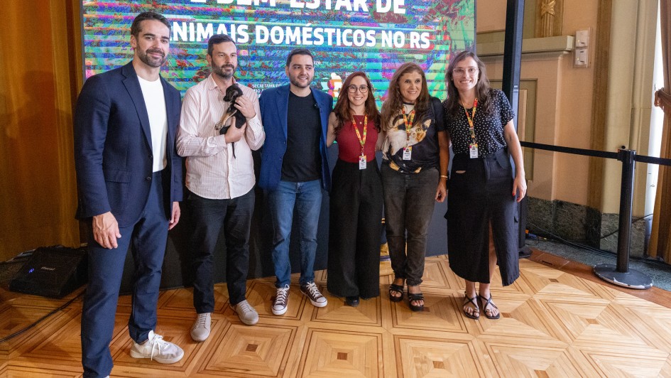 A fotografia colorida registra um momento de intera&ccedil;&atilde;o entre autoridades e a comunidade durante um evento oficial voltado ao bem-estar animal.

Detalhes da cena:

Os participantes: No centro da imagem, o Governador Eduardo Leite, vestindo uma camisa social azul clara e cal&ccedil;a escura, est&aacute; levemente agachado para interagir com um cachorro. &Agrave; esquerda, uma mulher de blusa rosa e &oacute;culos sorri enquanto observa a cena. Ao redor deles, outras pessoas acompanham o momento em um ambiente de celebra&ccedil;&atilde;o.

O animal: Um cachorro de m&eacute;dio porte, com pelagem curta e amarelada (semelhante a um Labrador ou vira-lata), est&aacute; em p&eacute; &agrave; frente do governador. O animal parece calmo e recebe aten&ccedil;&atilde;o das autoridades.

O ambiente: O evento ocorre em um espa&ccedil;o amplo com ilumina&ccedil;&atilde;o natural. Ao fundo, &eacute; poss&iacute;vel ver estruturas que sugerem um parque ou &aacute;rea externa coberta, com &aacute;rvores e folhagens vis&iacute;veis ao longe. H&aacute; tamb&eacute;m um banner ou painel com identidades visuais do governo do Estado.

Composi&ccedil;&atilde;o e Ilumina&ccedil;&atilde;o: A foto captura o grupo da cintura para cima, com foco central na intera&ccedil;&atilde;o entre o governador e o c&atilde;o. A luz do dia &eacute; clara, destacando as express&otilde;es de satisfa&ccedil;&atilde;o dos presentes e refor&ccedil;ando o car&aacute;ter p&uacute;blico e amig&aacute;vel da agenda.