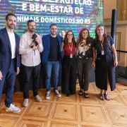 A fotografia colorida registra um momento de intera&ccedil;&atilde;o entre autoridades e a comunidade durante um evento oficial voltado ao bem-estar animal.

Detalhes da cena:

Os participantes: No centro da imagem, o Governador Eduardo Leite, vestindo uma camisa social azul clara e cal&ccedil;a escura, est&aacute; levemente agachado para interagir com um cachorro. &Agrave; esquerda, uma mulher de blusa rosa e &oacute;culos sorri enquanto observa a cena. Ao redor deles, outras pessoas acompanham o momento em um ambiente de celebra&ccedil;&atilde;o.

O animal: Um cachorro de m&eacute;dio porte, com pelagem curta e amarelada (semelhante a um Labrador ou vira-lata), est&aacute; em p&eacute; &agrave; frente do governador. O animal parece calmo e recebe aten&ccedil;&atilde;o das autoridades.

O ambiente: O evento ocorre em um espa&ccedil;o amplo com ilumina&ccedil;&atilde;o natural. Ao fundo, &eacute; poss&iacute;vel ver estruturas que sugerem um parque ou &aacute;rea externa coberta, com &aacute;rvores e folhagens vis&iacute;veis ao longe. H&aacute; tamb&eacute;m um banner ou painel com identidades visuais do governo do Estado.

Composi&ccedil;&atilde;o e Ilumina&ccedil;&atilde;o: A foto captura o grupo da cintura para cima, com foco central na intera&ccedil;&atilde;o entre o governador e o c&atilde;o. A luz do dia &eacute; clara, destacando as express&otilde;es de satisfa&ccedil;&atilde;o dos presentes e refor&ccedil;ando o car&aacute;ter p&uacute;blico e amig&aacute;vel da agenda.