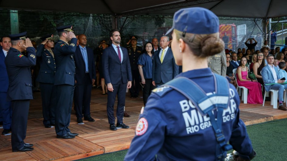 Foto mostra o governador Eduardo Leite no centro da foto. Militares fazem contin&ecirc;ncia para ele. Em primeiro plano, de costas para a c&acirc;mera, uma bombeiro militar fardada, tamb&eacute;m est&aacute; direcionada para ele.