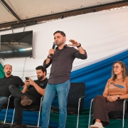 Quatro pessoasl em um palco, tr&ecirc;s sentadas e uma em p&eacute; falando ao microfone para pessoas &agrave; frente que n&atilde;o aparecem na foto