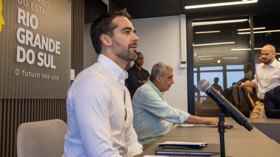 Dois homens sentados com os bra&ccedil;os apoiados em uma grande mesa, &agrave; frente de um painel com os dizeres: Governo do Estado do Rio Grande do Sul O Futuro nos Une. Um terceiro homem em p&eacute; olha para ambos 