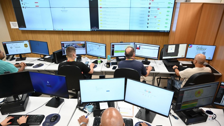 Sala de monitoramento com v&aacute;rias esta&ccedil;&otilde;es de trabalho alinhadas em mesas brancas. Cada esta&ccedil;&atilde;o possui dois monitores exibindo mapas, gr&aacute;ficos, listas de ocorr&ecirc;ncias e pain&eacute;is de controle digital.