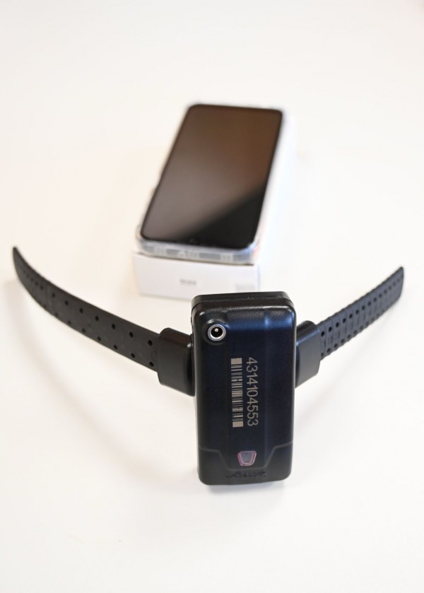 Dispositivo de monitoramento eletrônico com formato retangular e pulseira de borracha perfurada, apoiado sobre uma superfície branca. Ao fundo, há um telefone celular apoiado sobre a respectiva caixa, levemente desfocado, compondo um arranjo de demonstração do sistema.