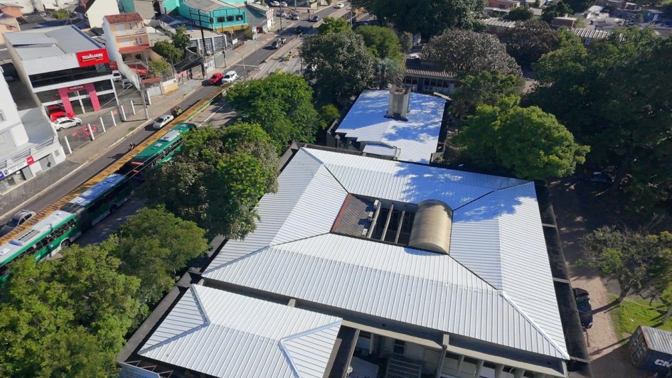 A fotografia colorida mostra um grande grupo de pessoas, composto por adultos e crian&ccedil;as, reunido em um p&aacute;tio escolar coberto para uma celebra&ccedil;&atilde;o coletiva.

Detalhes da cena:

O Grupo: O grupo &eacute; diversificado e ocupa quase todo o enquadramento. &Agrave; frente, crian&ccedil;as vestindo uniformes escolares (camisetas brancas ou verdes com detalhes em amarelo e azul) sorriem e acenam com as m&atilde;os para o alto. Atr&aacute;s delas, adultos &mdash; incluindo professores, gestores e possivelmente autoridades &mdash; tamb&eacute;m acenam e celebram.

Destaques: No centro do grupo, uma mulher segura um documento oficial emoldurado ou em uma pasta, indicando a formaliza&ccedil;&atilde;o de uma entrega ou projeto. Logo &agrave; frente, uma mulher agachada segura um microfone e sorri para a c&acirc;mera, sugerindo que ela coordenava o evento.

O Ambiente: O local &eacute; um p&aacute;tio coberto com p&eacute;-direito alto. A estrutura possui pilares e paredes pintadas em um tom de azul vibrante. O teto &eacute; forrado com placas brancas e possui lumin&aacute;rias embutidas. Ao fundo, as paredes s&atilde;o azuis e h&aacute; placas informativas fixadas.

Ilumina&ccedil;&atilde;o e Clima: A ilumina&ccedil;&atilde;o &eacute; natural e abundante, vinda das laterais abertas do p&aacute;tio. O clima da imagem &eacute; de grande entusiasmo, alegria e conquista compartilhada entre a comunidade escolar e os gestores presentes.

Composi&ccedil;&atilde;o: A foto &eacute; tirada de um &acirc;ngulo frontal e ligeiramente elevado, o que permite visualizar a profundidade do grupo e a express&atilde;o de felicidade de quase todos os participantes.