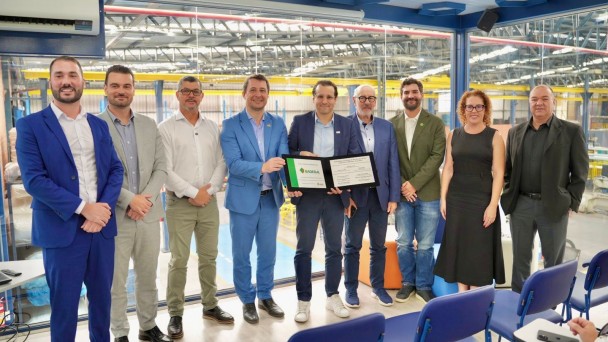 Um grupo de nove pessoas posa em p&eacute; lado a lado para a foto em um ambiente industrial. Ao centro, dois homens seguram e exibem uma placa de reconhecimento. 